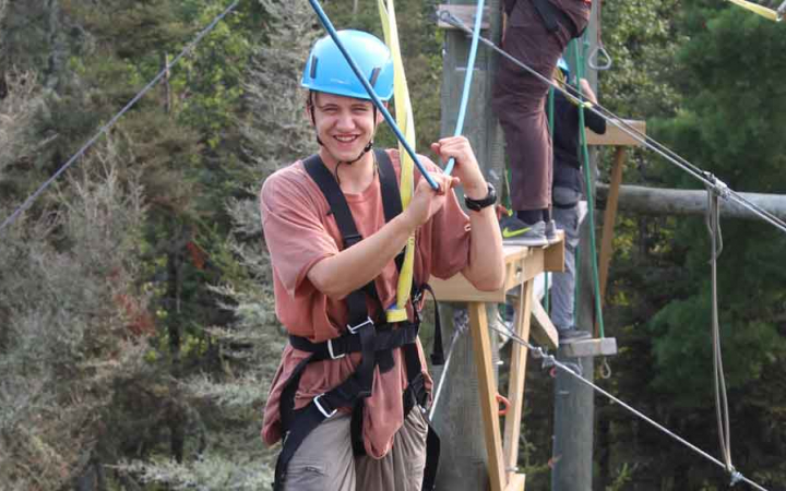 A person wearing safety gear and secured by ropes balances on an obstacle on a high ropes course. 